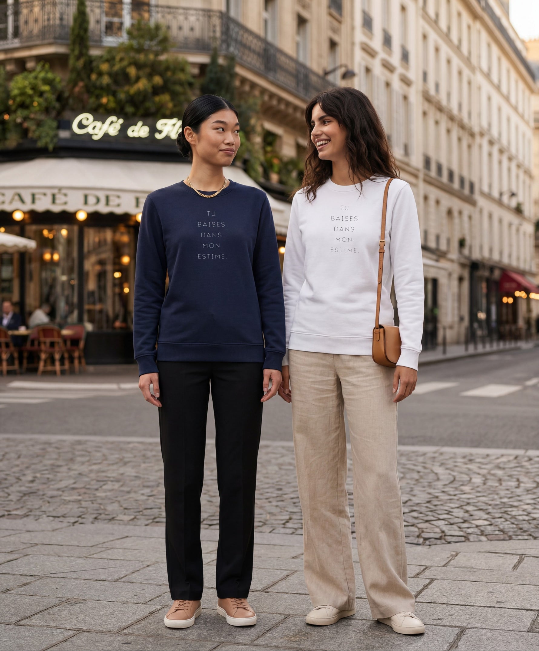 Deux femmes portant des sweatshirts Salut Fred « Tu baises dans mon estime. », l’une en bleu marine et l’autre en blanc, dans une rue parisienne devant un café parisien classique