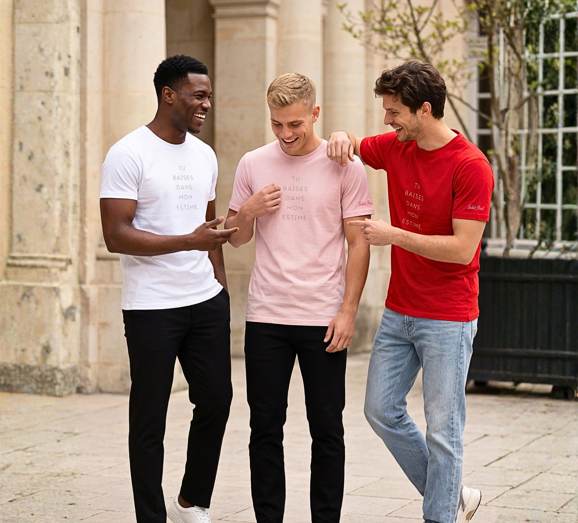 Groupe lifestyle de trois hommes portant les t-shirts classiques « Tu baises dans mon estime. » en blanc, rose poudré et rouge avec texte imprimé centré sur la poitrine et logo Salut Fred sur la manche gauche, dans une cour parisienne lumineuse.