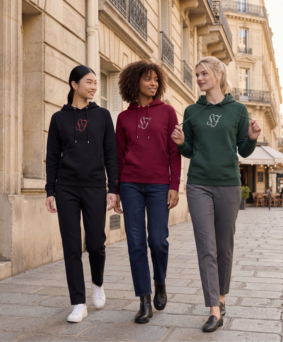 Vue de face de trois femmes portant une sélection de hoodies SF Monogramme Salut Fred en bordeaux, noir et vert profond dans une rue parisienne bordée d’immeubles haussmanniens