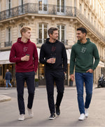 Vue de face de trois hommes portant une sélection de hoodies SF Monogramme Salut Fred en bordeaux, noir et vert profond dans une rue parisienne bordée d’immeubles haussmanniens