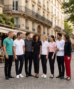 Groupe d’hommes et de femmes portant la collection « Thérap(e)ute. À votre service » de Salut Fred dans une rue parisienne élégante, avec t-shirts classiques, polos et t-shirts manches longues en jade, blanc, gris graphite, noir et rose poudré, tous avec motif brodé sur la poitrine gauche et logos Salut Fred brodés sur les manches ou poignets