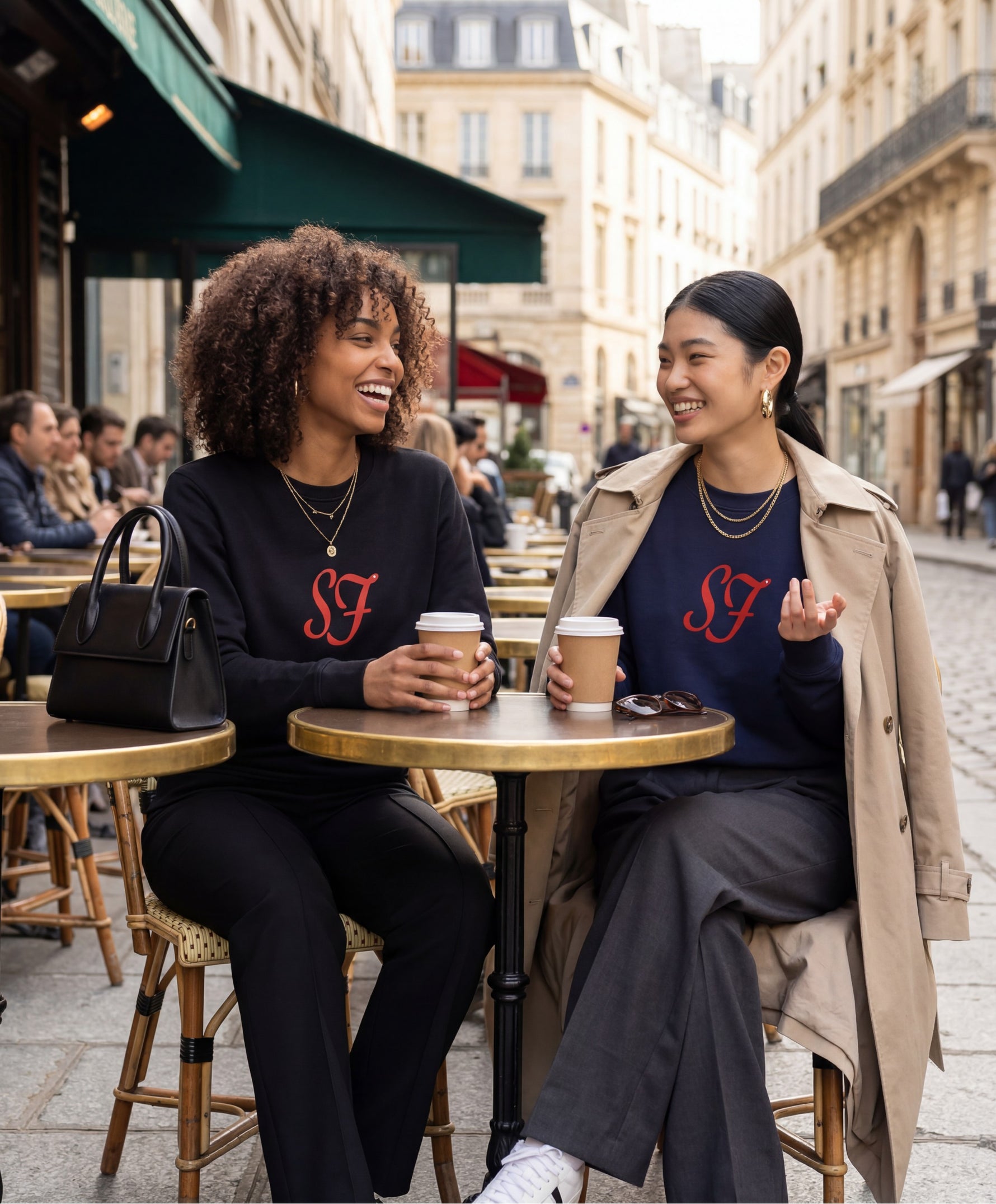 Vue de face de deux femmes portant les sweatshirts SF Monogramme Salut Fred en noir et bleu marine avec monogramme SF imprimé au centre de la poitrine en terrasse de café dans une rue parisienne animée