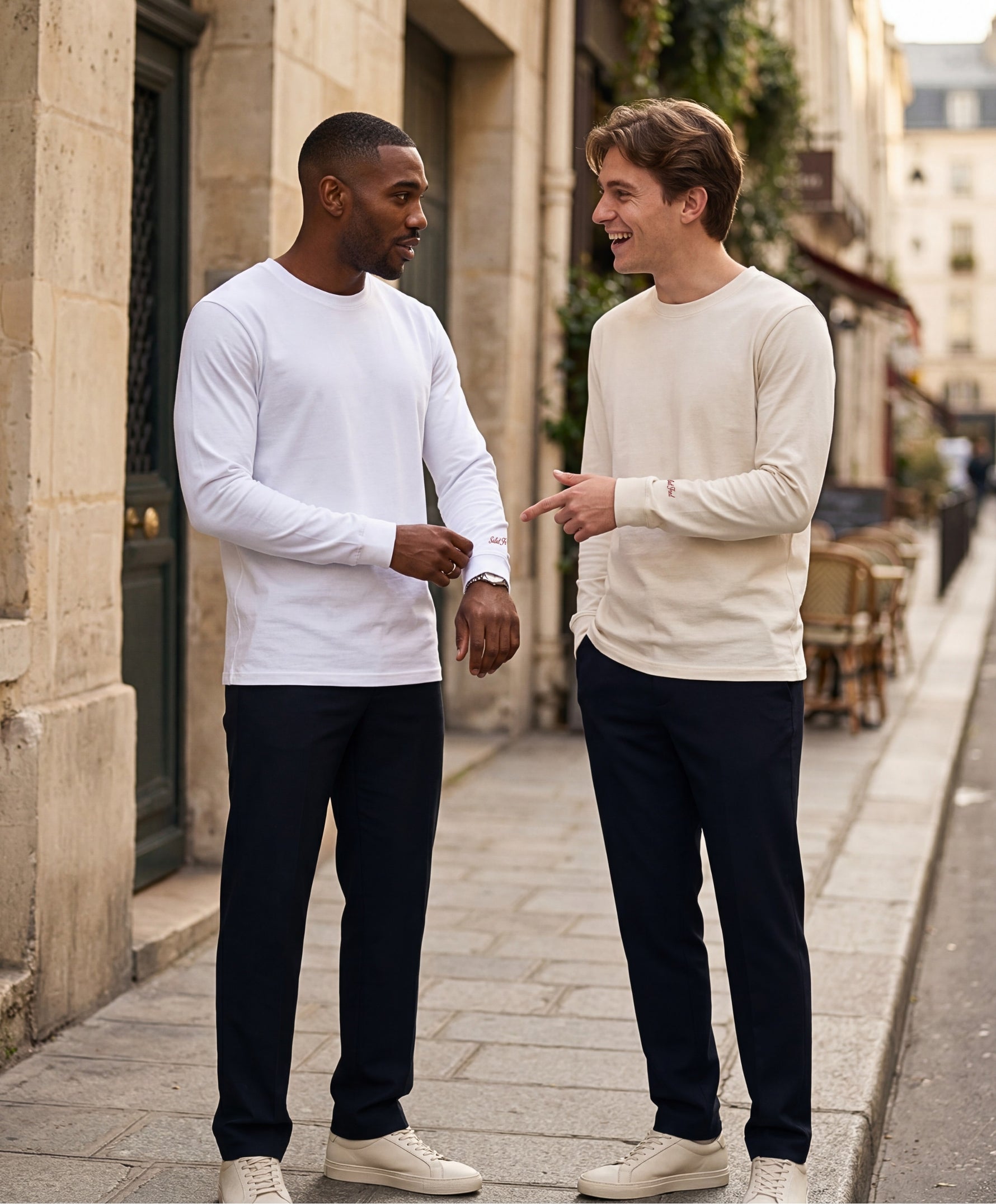 Deux hommes portant des t-shirts à manches longues Salut Fred blanc et écru avec broderie rouge sur le poignet gauche, dans une rue parisienne lumineuse aux façades haussmanniennes élégantes.