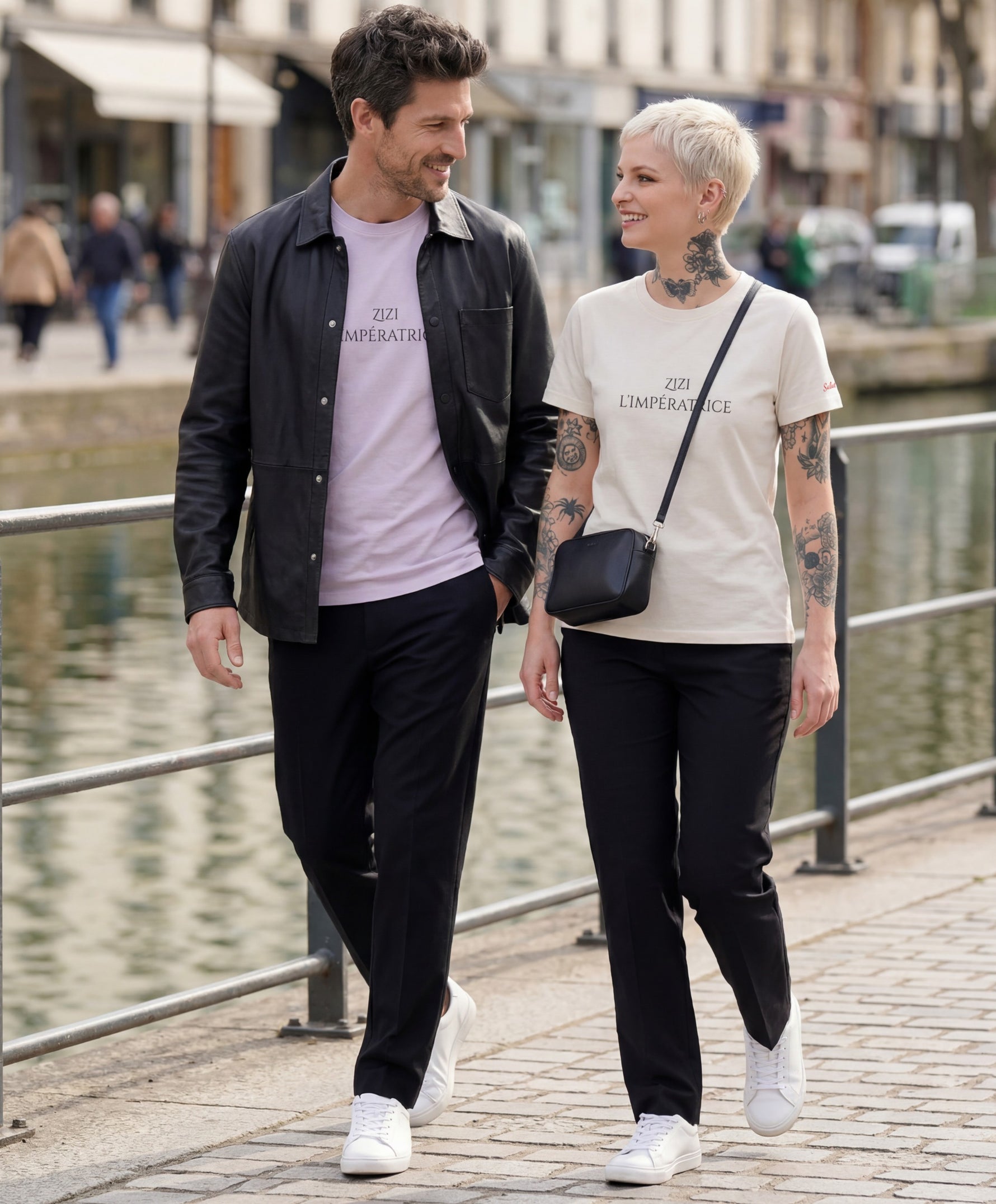 Vue lifestyle d’un homme portant le t-shirt classique lilas « Zizi l’Impératrice » et d’une femme portant le t-shirt classique écru « Zizi l’Impératrice », avec texte imprimé noir centré sur la poitrine et logo Salut Fred sur la manche gauche, en promenade au bord d’un canal parisien.