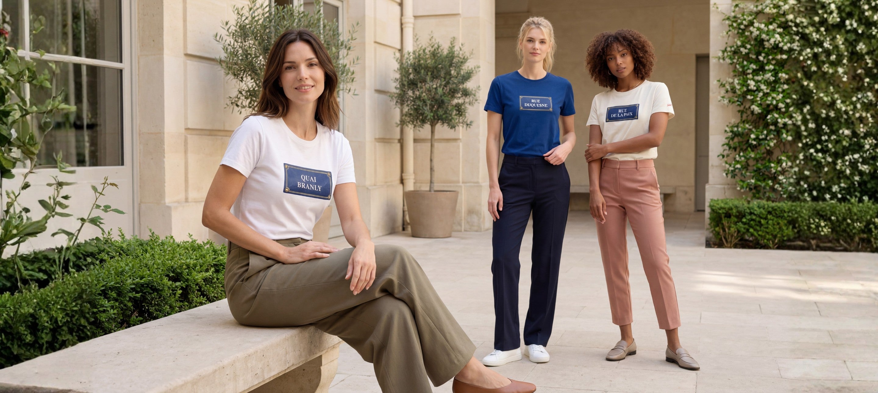 Ligne de t-shirts classiques Salut Fred en blanc, bleu Paris et écru, portés par trois femmes dans une cour parisienne élégante avec imprimés style plaques de rue françaises.