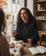 Vue de face d’une femme portant un hoodie noir avec monogramme SF brodé en rouge, assise dans un café chaleureux autour d’un café et de pâtisseries
