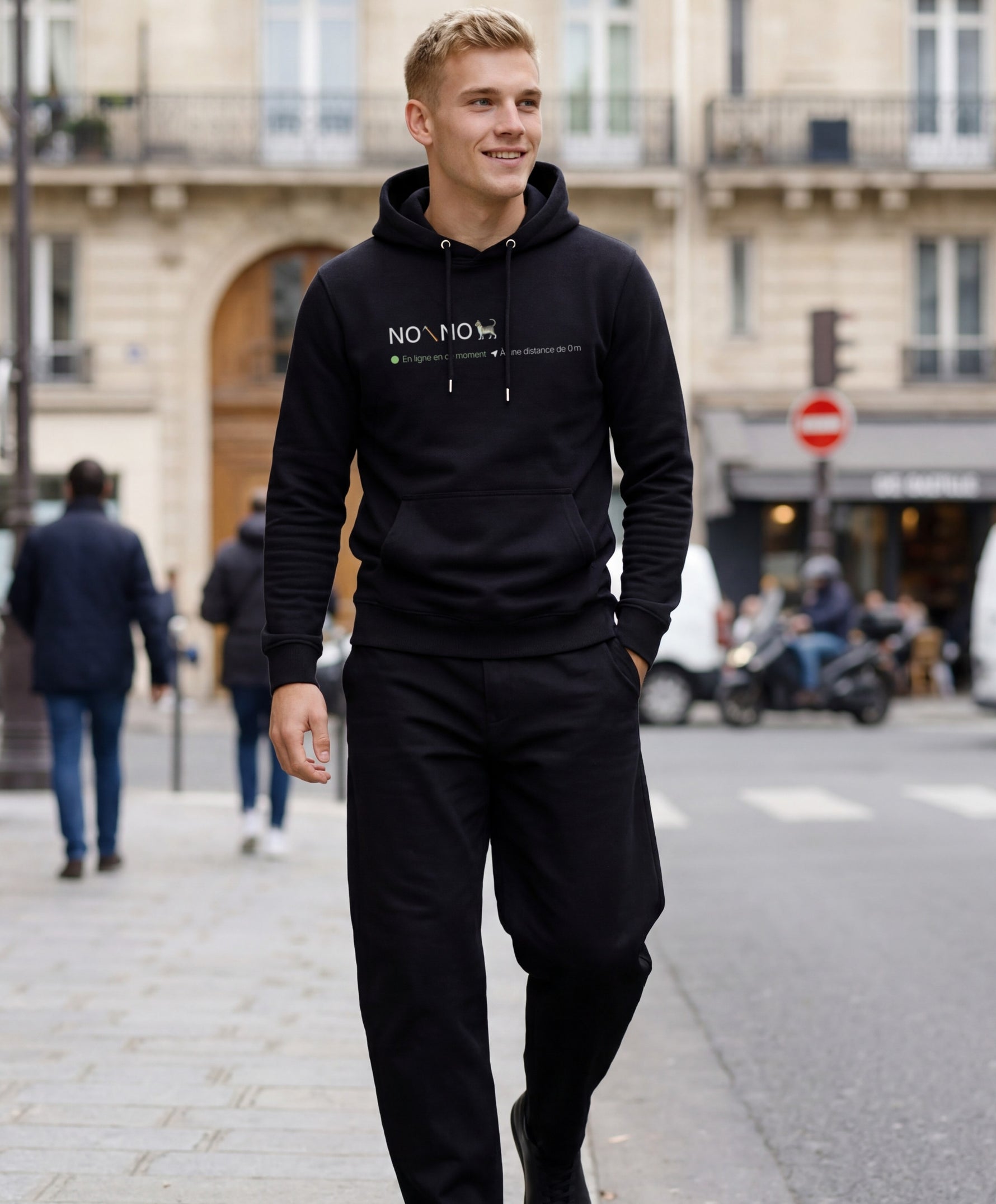 Vue de face d’un homme portant un hoodie noir Salut Fred avec motif « No pic No Chat(te) », où « pic » et « Chat(te) » sont représentés par des emojis, dans une rue parisienne élégante