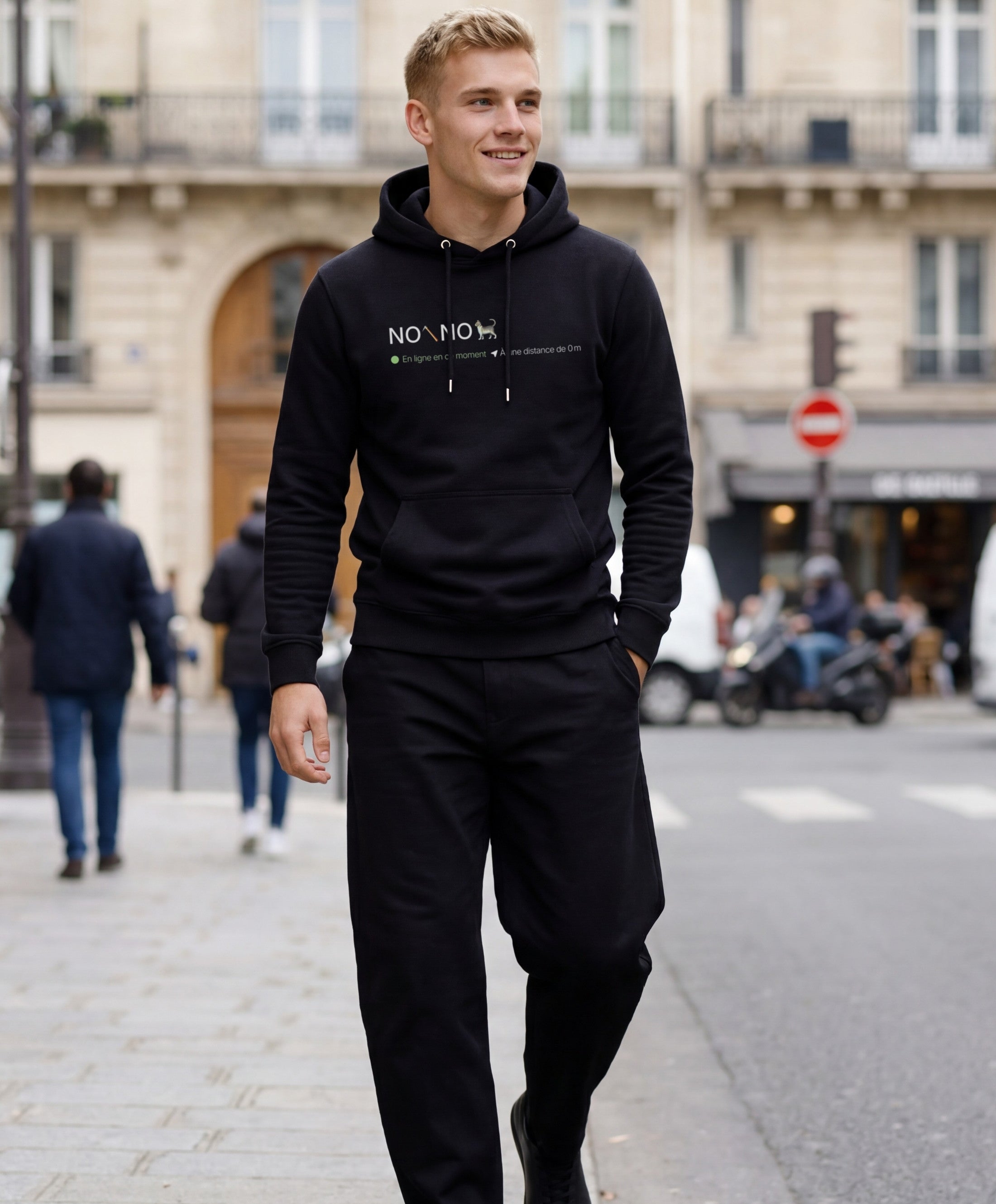 Vue de face d’un homme portant un hoodie noir Salut Fred avec motif « No pic No Chat(te) », où « pic » et « Chat(te) » sont représentés par des emojis, dans une rue parisienne élégante