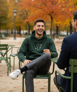 Vue de face d’un homme portant le hoodie vert profond Salut Fred avec monogramme SF blanc brodé au centre de la poitrine, assis dans un jardin parisien aux couleurs d’automne