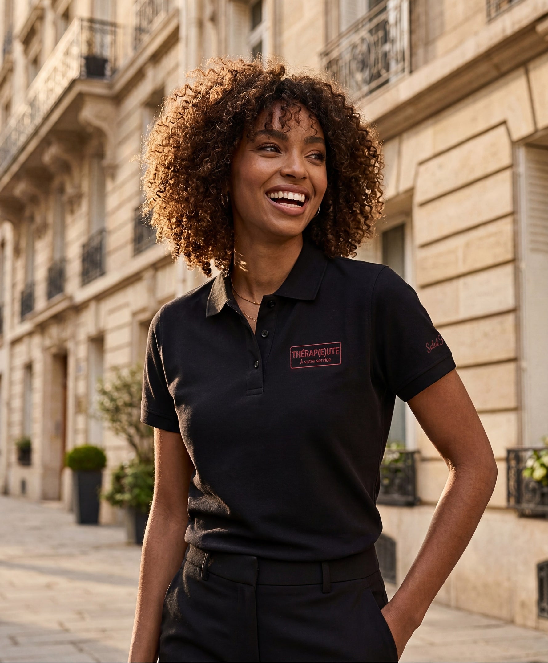 Vue de face d’une femme portant un polo noir Salut Fred avec broderie « Thérap(e)ute. À votre service » sur la poitrine et Salut Fred brodé sur la manche gauche dans une rue parisienne élégante