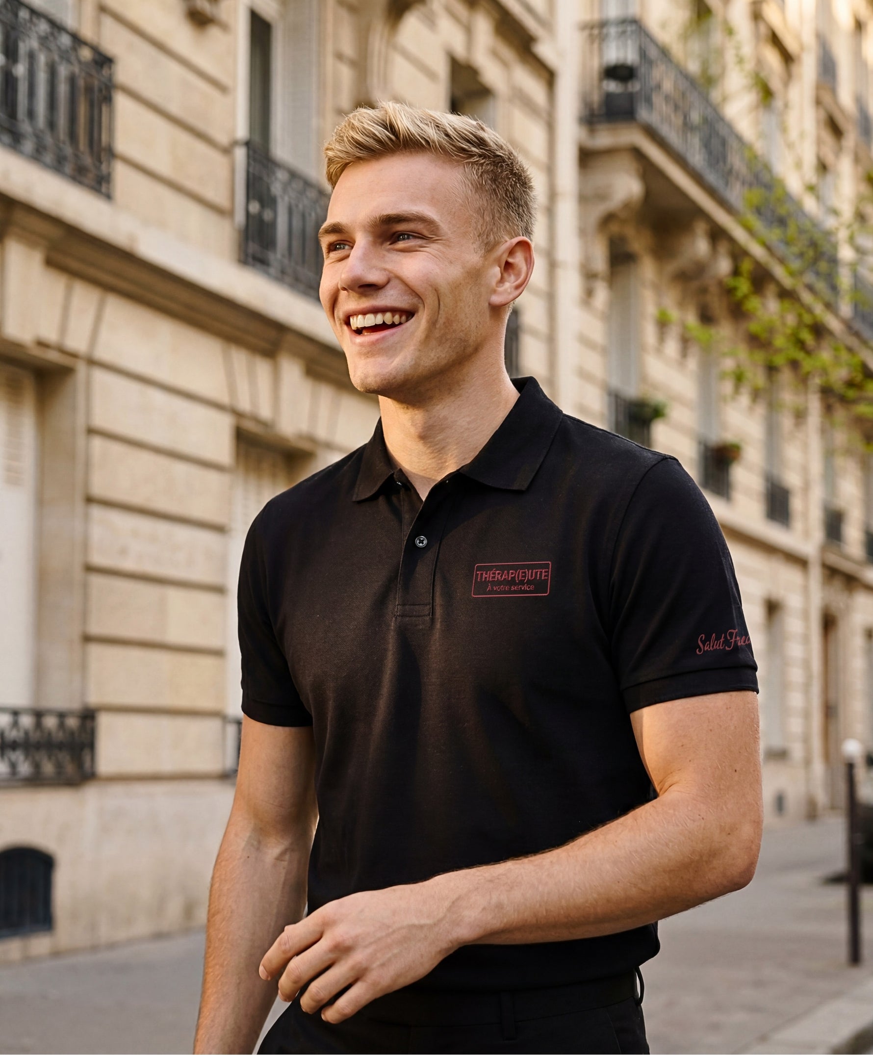 Vue de face d’un homme portant un polo noir Salut Fred avec broderie « Thérap(e)ute. À votre service » sur la poitrine et Salut Fred brodé sur la manche gauche dans une rue parisienne élégante