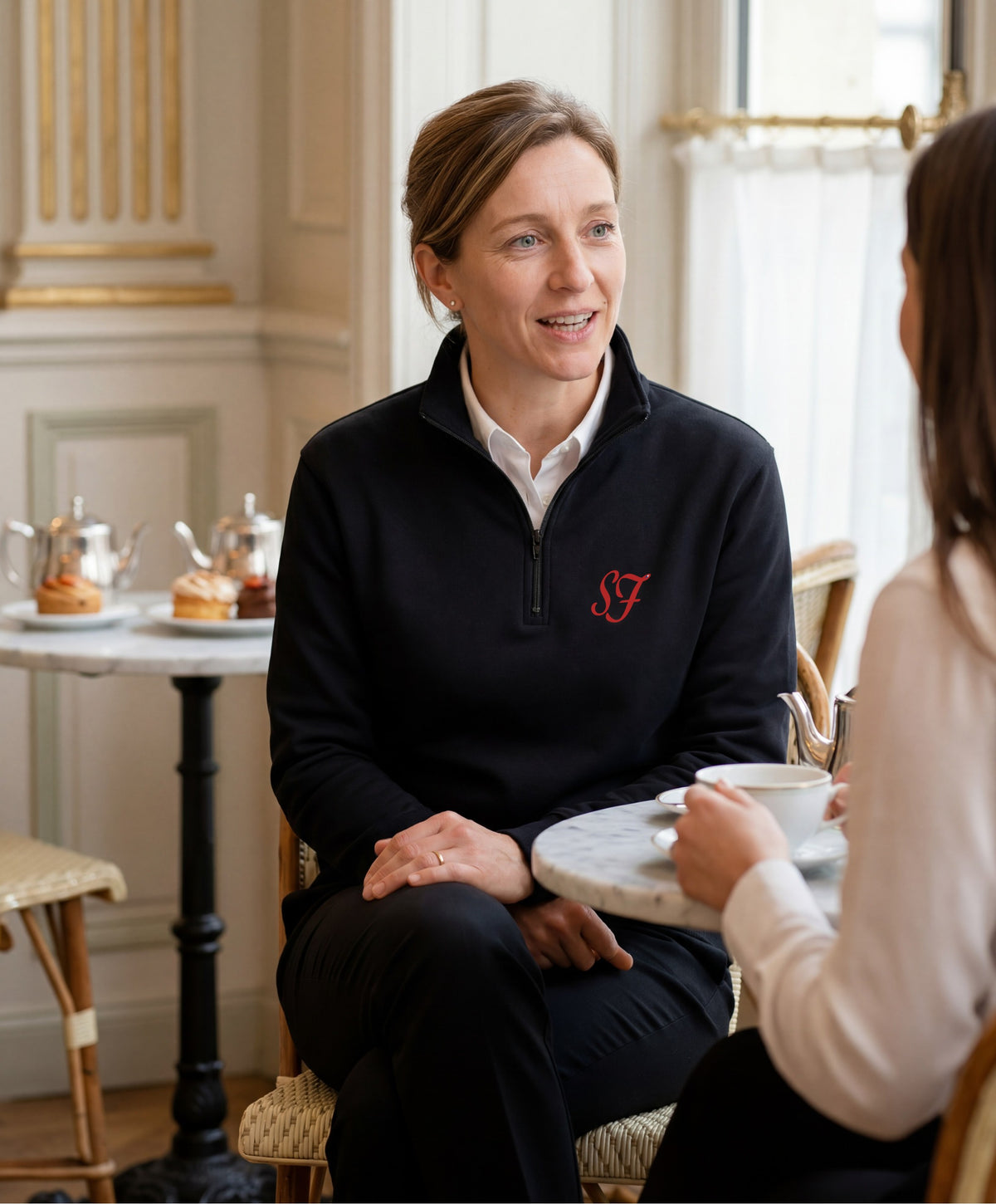 Vue de face d’une femme portant le sweatshirt à col zippé 1/4 noir SF Monogramme Salut Fred avec monogramme SF imprimé sur la poitrine dans un salon de thé parisien lumineux
