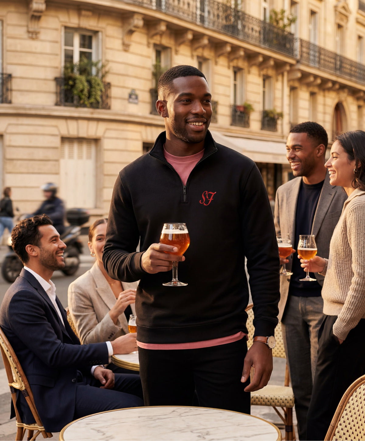 Vue de face d’un homme portant le sweatshirt à col zippé 1/4 noir SF Monogramme Salut Fred avec monogramme SF imprimé sur la poitrine en terrasse de café parisienne lors d’un apéritif entre amis