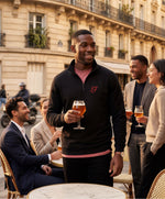 Vue de face d’un homme portant le sweatshirt à col zippé 1/4 noir SF Monogramme Salut Fred avec monogramme SF imprimé sur la poitrine en terrasse de café parisienne lors d’un apéritif entre amis