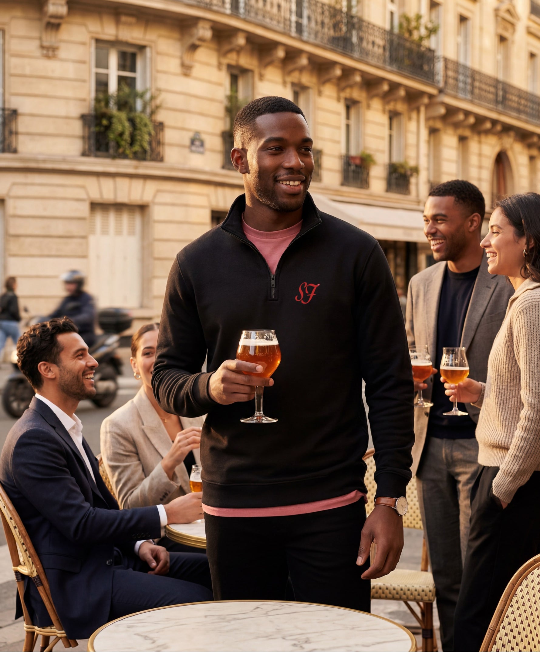 Vue de face d’un homme portant le sweatshirt à col zippé 1/4 noir SF Monogramme Salut Fred avec monogramme SF imprimé sur la poitrine en terrasse de café parisienne lors d’un apéritif entre amis