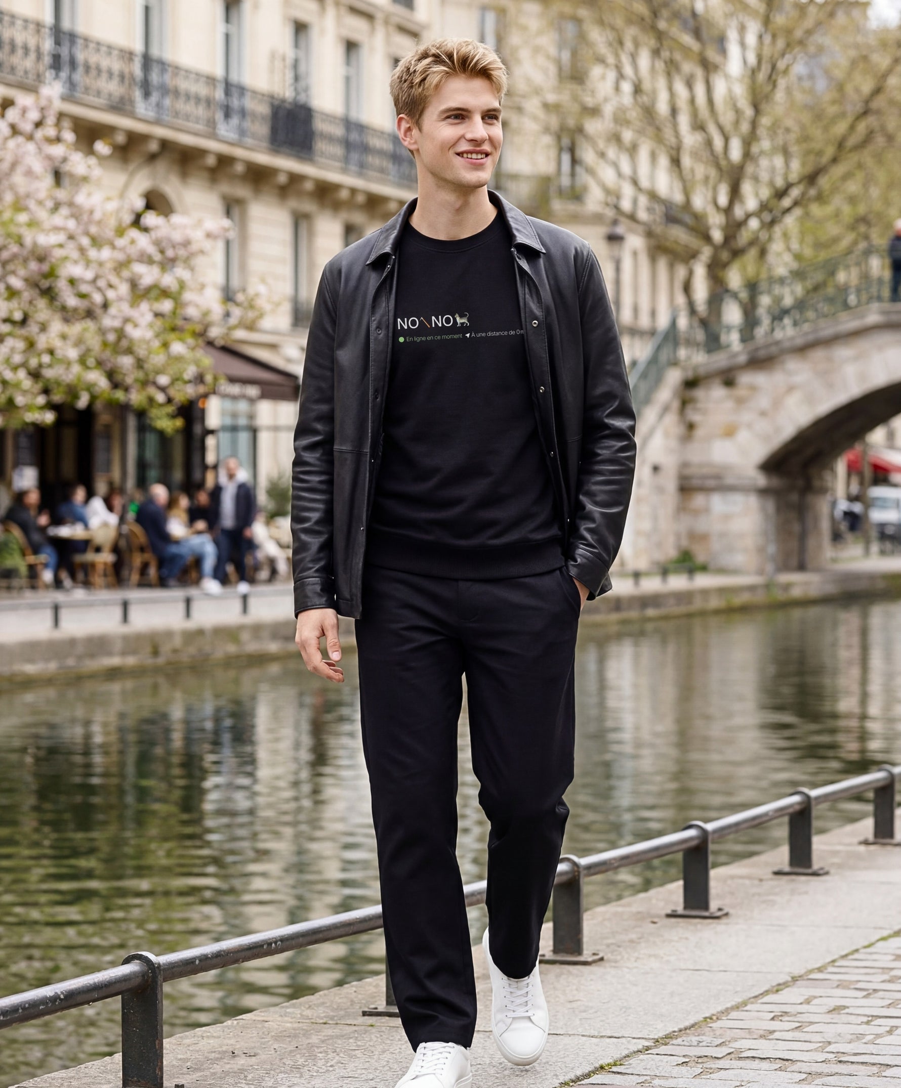 Vue de face d’un homme portant un sweatshirt noir Salut Fred avec motif « No pic No Chat(te) », où « pic » et « Chat(te) » sont représentés par des emojis, au bord d’un canal parisien