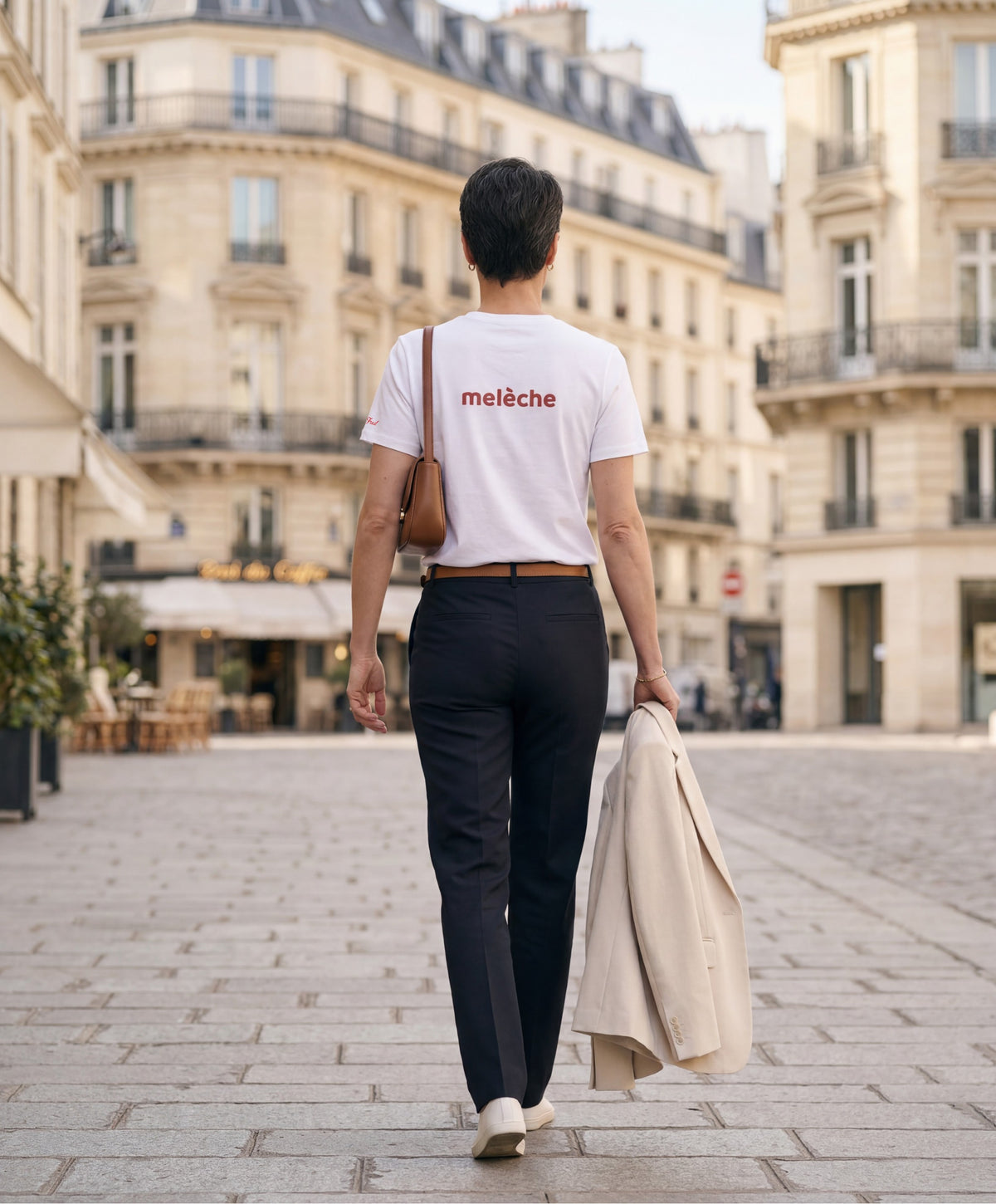 Vue de dos d’une femme portant un t-shirt classique blanc Salut Fred avec melèche imprimé au haut du dos dans une teinte melon pastel saumonée, pantalon noir ajusté, blazer écru à la main et sac en cuir camel dans une place parisienne élégante