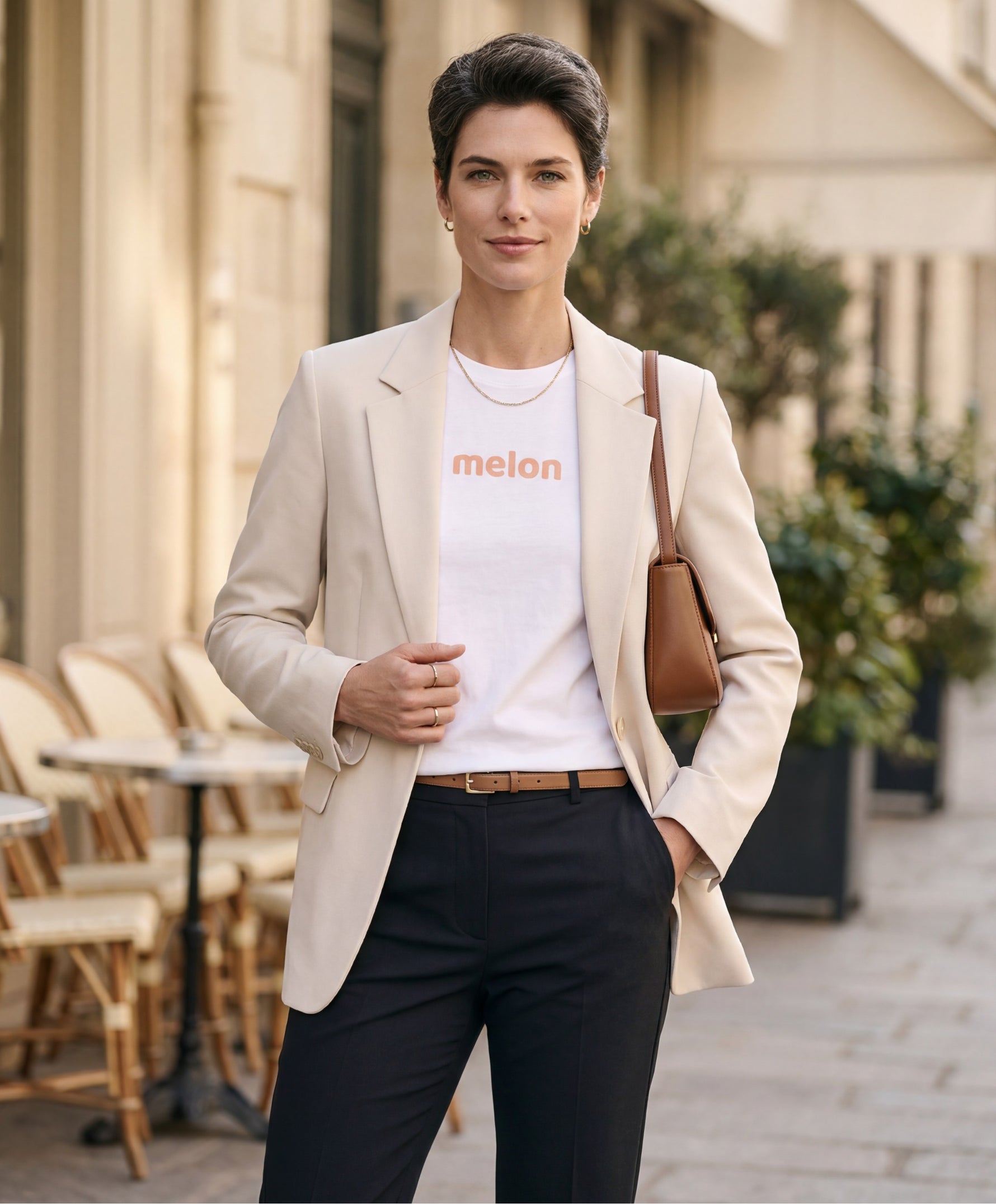 Femme portant un t-shirt classique blanc Salut Fred avec melon imprimé sur le devant dans une teinte melon pastel saumonée, blazer écru structuré, pantalon noir taille haute et sac en cuir camel dans une rue parisienne élégante
