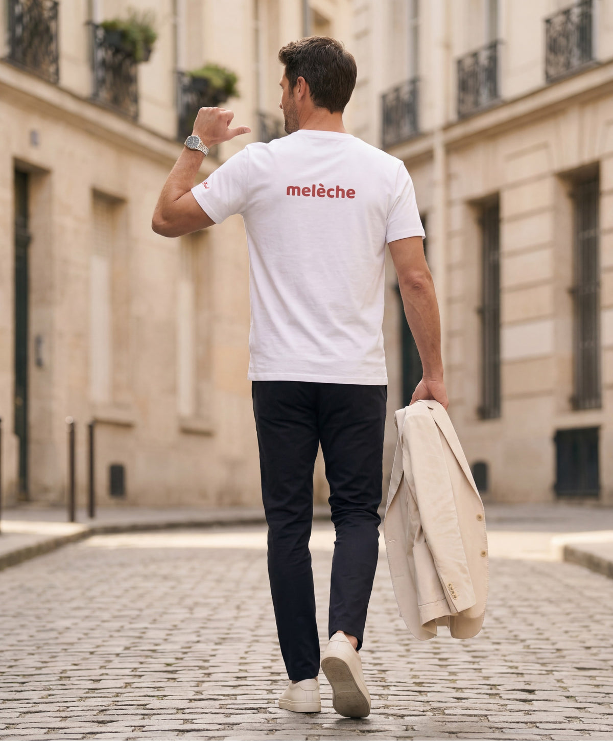 Vue de dos d’un homme portant un t-shirt classique blanc Salut Fred avec melèche imprimé au haut du dos dans une teinte melon pastel saumonée, pantalon bleu marine et blazer écru à la main dans une rue parisienne pavée