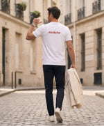 Vue de dos d’un homme portant un t-shirt classique blanc Salut Fred avec melèche imprimé au haut du dos dans une teinte melon pastel saumonée, pantalon bleu marine et blazer écru à la main dans une rue parisienne pavée