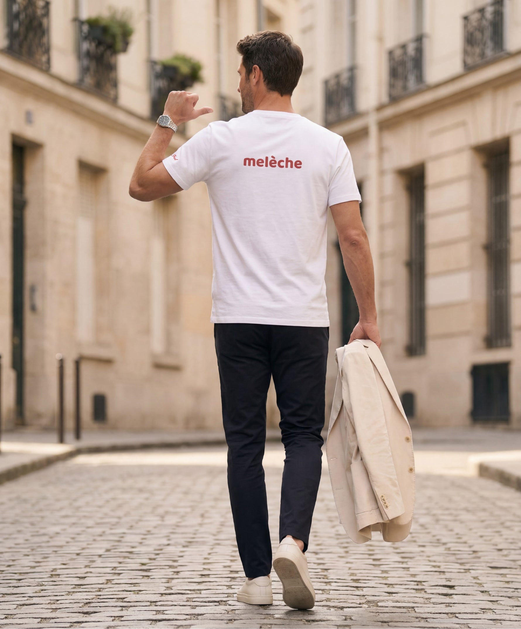 Vue de dos d’un homme portant un t-shirt classique blanc Salut Fred avec melèche imprimé au haut du dos dans une teinte melon pastel saumonée, pantalon bleu marine et blazer écru à la main dans une rue parisienne pavée