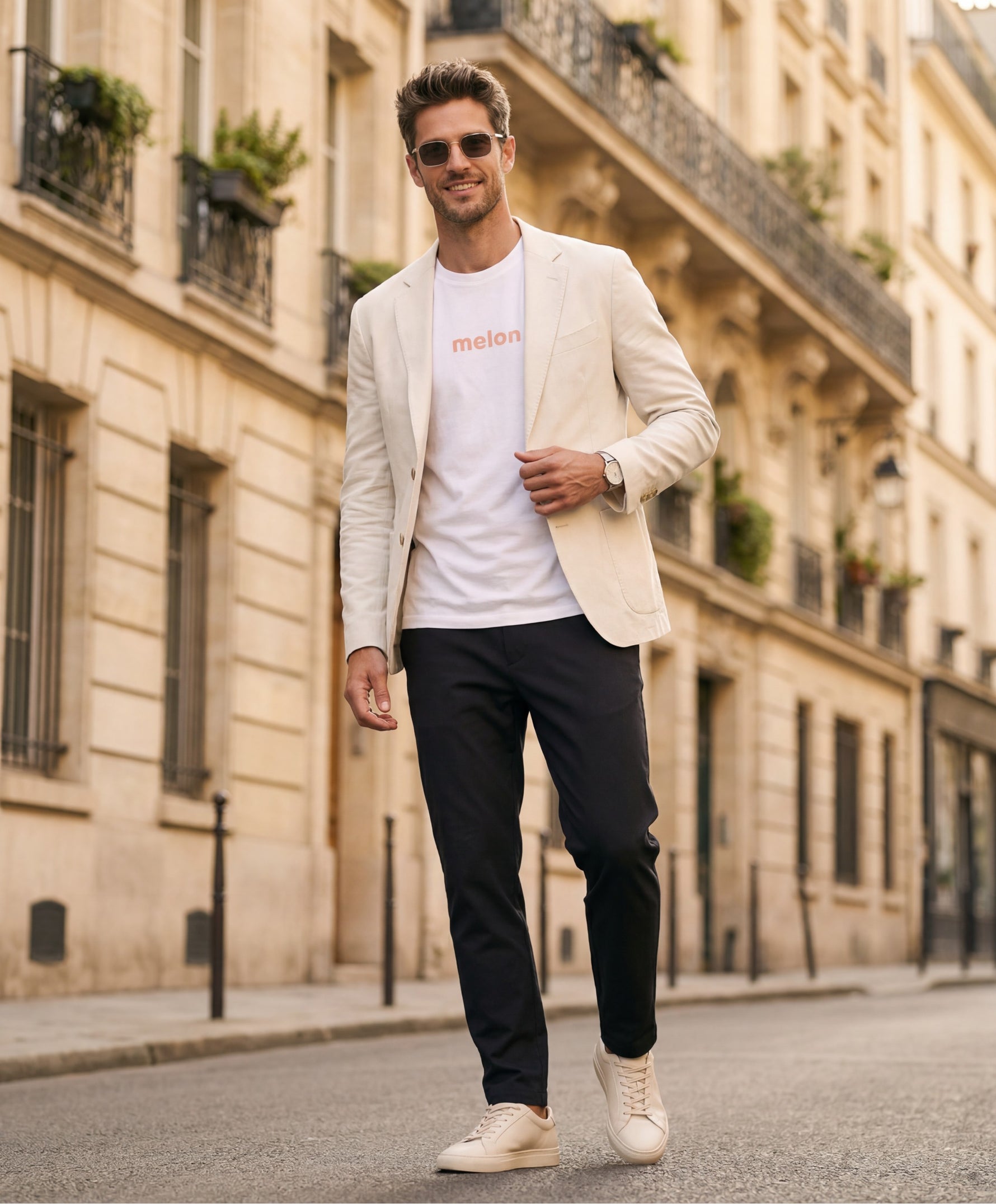 Vue de face d’un homme portant un t-shirt classique blanc Salut Fred avec melon imprimé au centre de la poitrine dans une teinte melon pastel saumonée, blazer écru, pantalon bleu marine et sneakers écrues dans une rue parisienne élégante