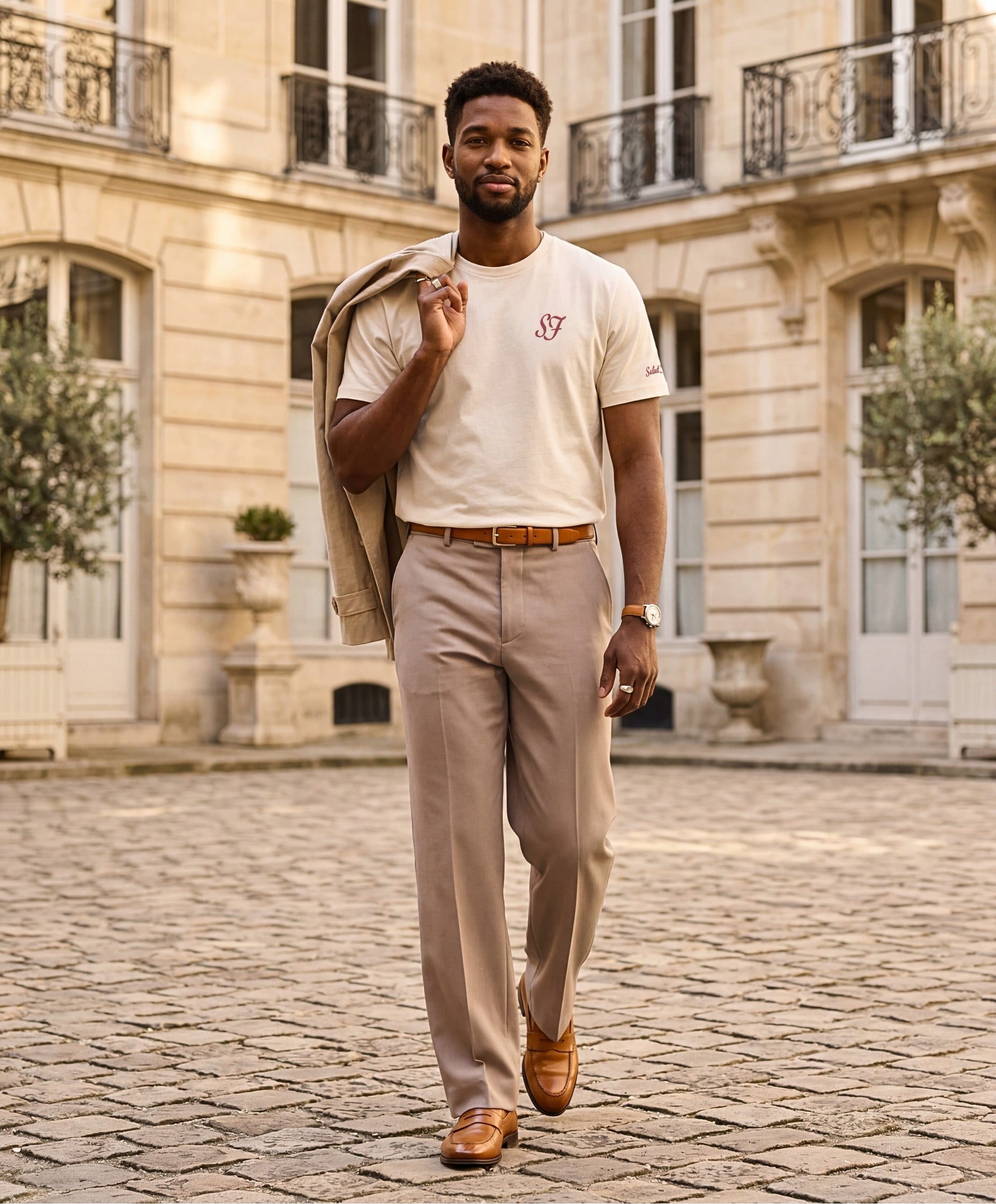 Vue de face d’un homme portant un t-shirt classique écru Salut Fred avec monogramme SF brodé sur la poitrine gauche, broderie Salut Fred sur la manche gauche, pantalon beige habillé, ceinture camel et mocassins camel dans une cour parisienne élégante