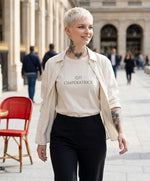 Vue de face d’une femme portant le t-shirt classique écru « Zizi l’Impératrice » avec texte imprimé noir centré sur la poitrine et logo Salut Fred sur la manche gauche, associé à une surchemise écru dans une galerie parisienne lumineuse.