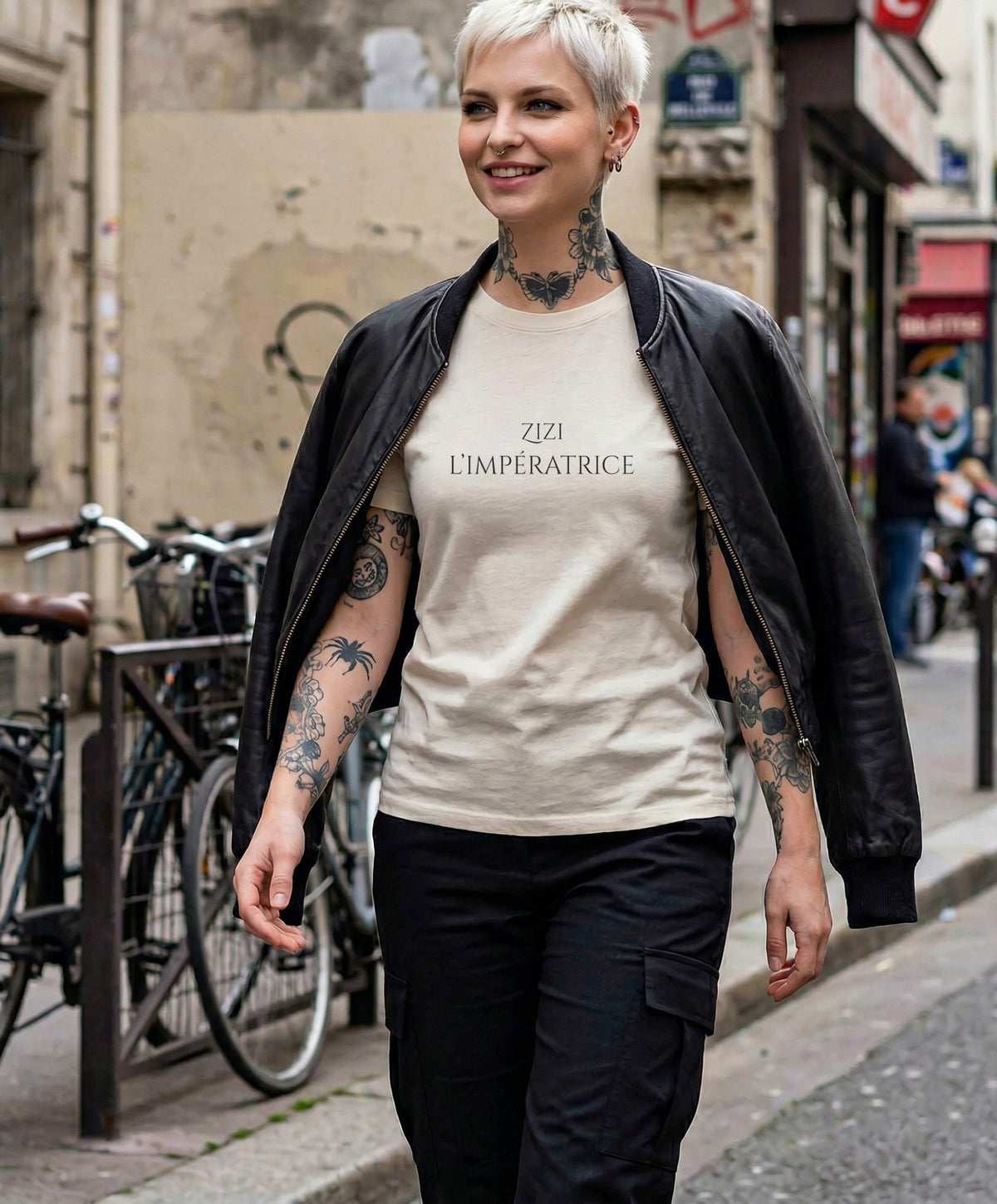 Vue de face d’une femme portant le t-shirt classique écru « Zizi l’Impératrice » avec texte imprimé noir centré sur la poitrine et logo Salut Fred sur la manche gauche, associé à un bomber noir dans une rue parisienne au style urbain.