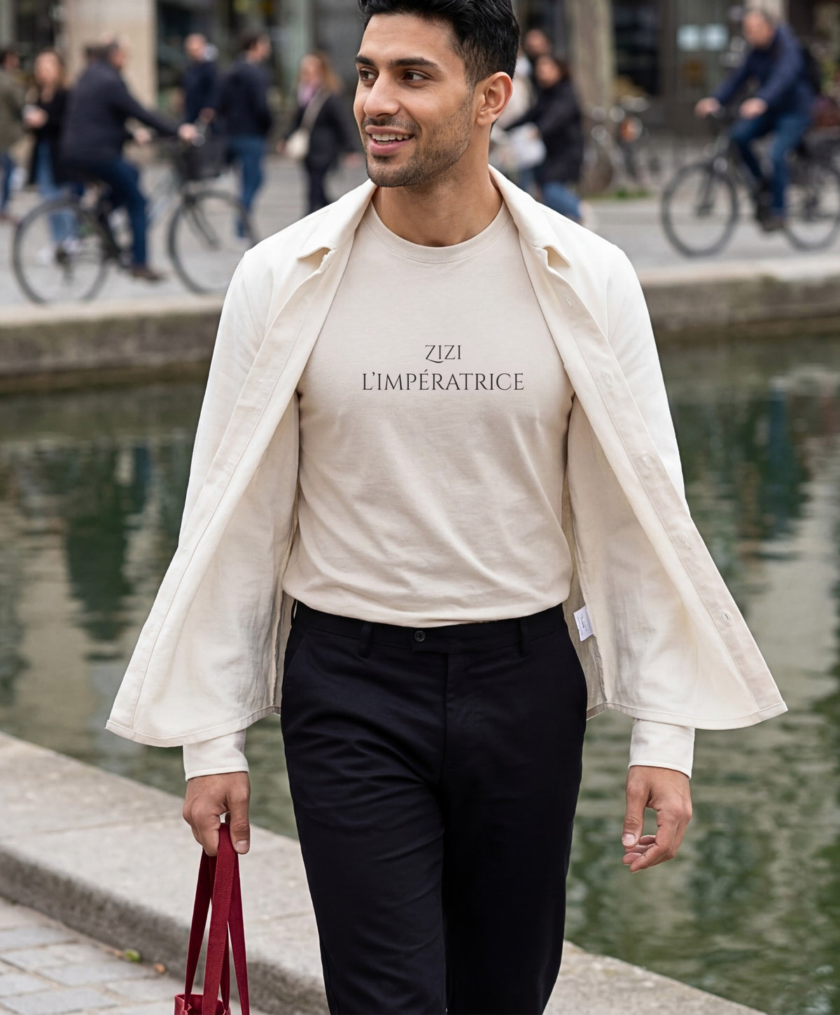 Vue de face d’un homme portant le t-shirt classique écru « Zizi l’Impératrice » avec texte imprimé noir centré sur la poitrine et logo Salut Fred sur la manche gauche, associé à une surchemise écru au bord d’un canal urbain.