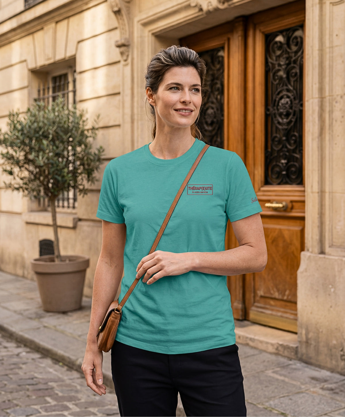 Femme portant un t-shirt classique jade Salut Fred avec broderie « Thérap(e)ute À votre service » sur la poitrine gauche et broderie Salut Fred sur la manche gauche, dans une rue parisienne devant une porte d’immeuble haussmannien