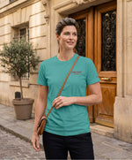 Femme portant un t-shirt classique jade Salut Fred avec broderie « Thérap(e)ute À votre service » sur la poitrine gauche et broderie Salut Fred sur la manche gauche, dans une rue parisienne devant une porte d’immeuble haussmannien