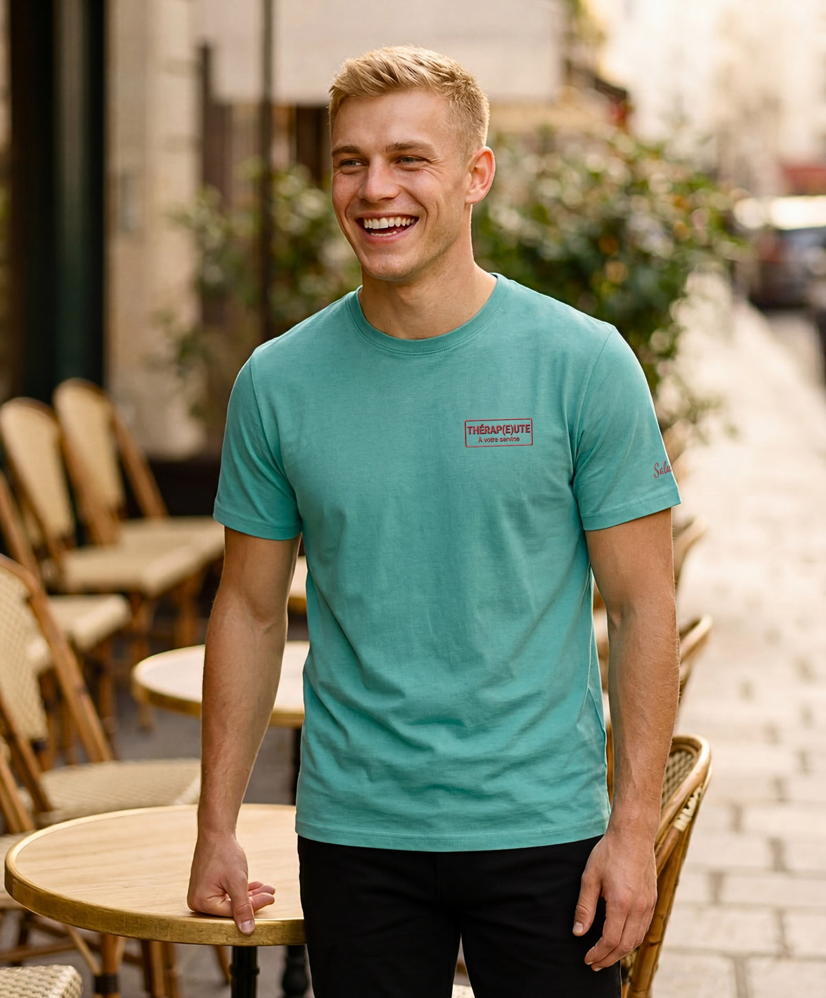 Homme portant un t-shirt classique jade Salut Fred avec broderie « Thérap(e)ute À votre service » sur la poitrine gauche et broderie Salut Fred sur la manche gauche, dans une rue parisienne devant une terrasse de café