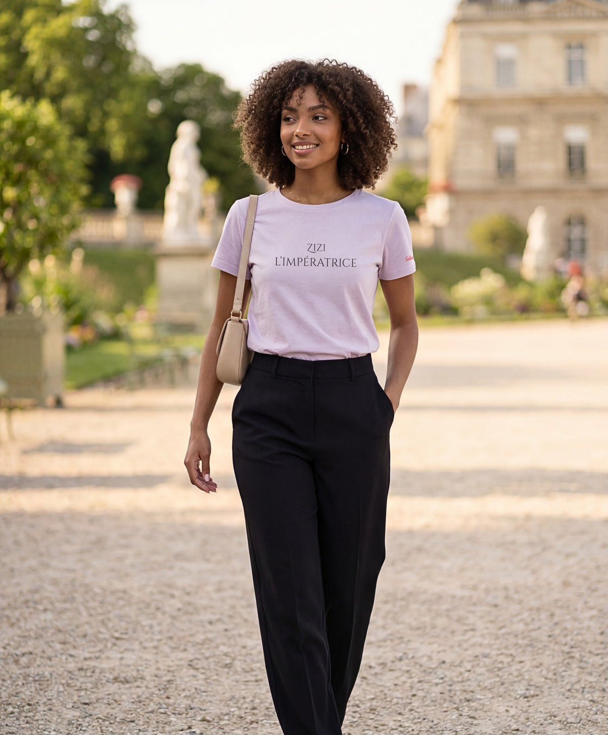 Vue de face d’une femme portant le t-shirt classique lilas « Zizi l’Impératrice » avec texte imprimé noir centré sur la poitrine et logo Salut Fred sur la manche gauche, dans un jardin parisien.