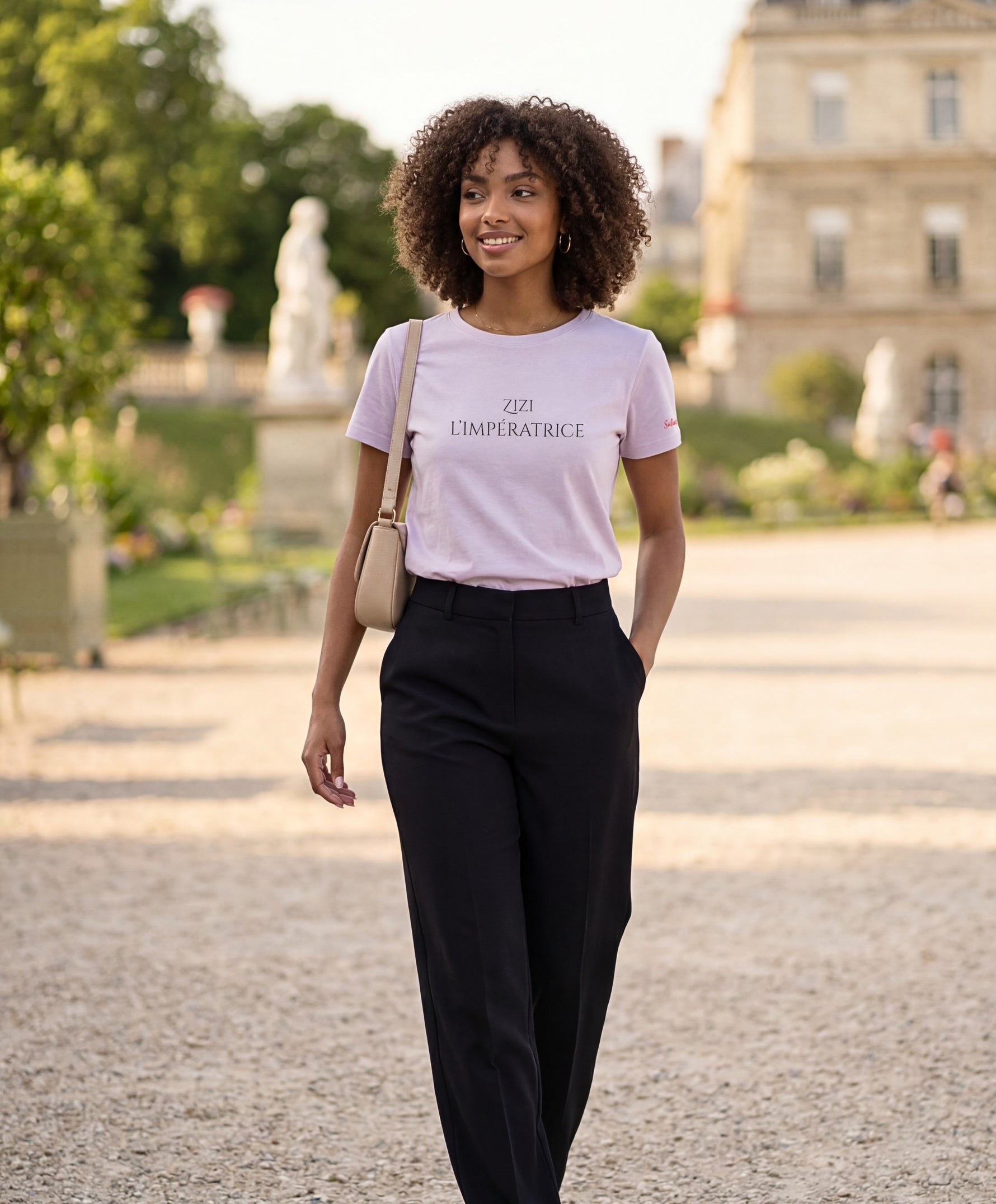 Vue de face d’une femme portant le t-shirt classique lilas « Zizi l’Impératrice » avec texte imprimé noir centré sur la poitrine et logo Salut Fred sur la manche gauche, dans un jardin parisien.