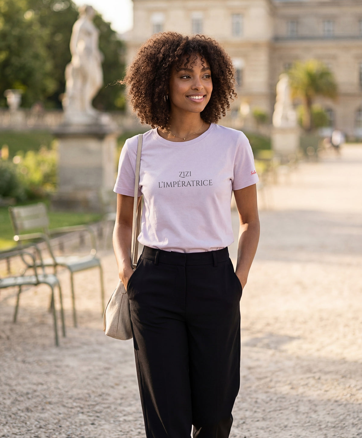 Vue de face d’une femme portant le t-shirt classique lilas « Zizi l’Impératrice » avec texte imprimé noir centré sur la poitrine et logo Salut Fred sur la manche gauche, dans un jardin parisien.