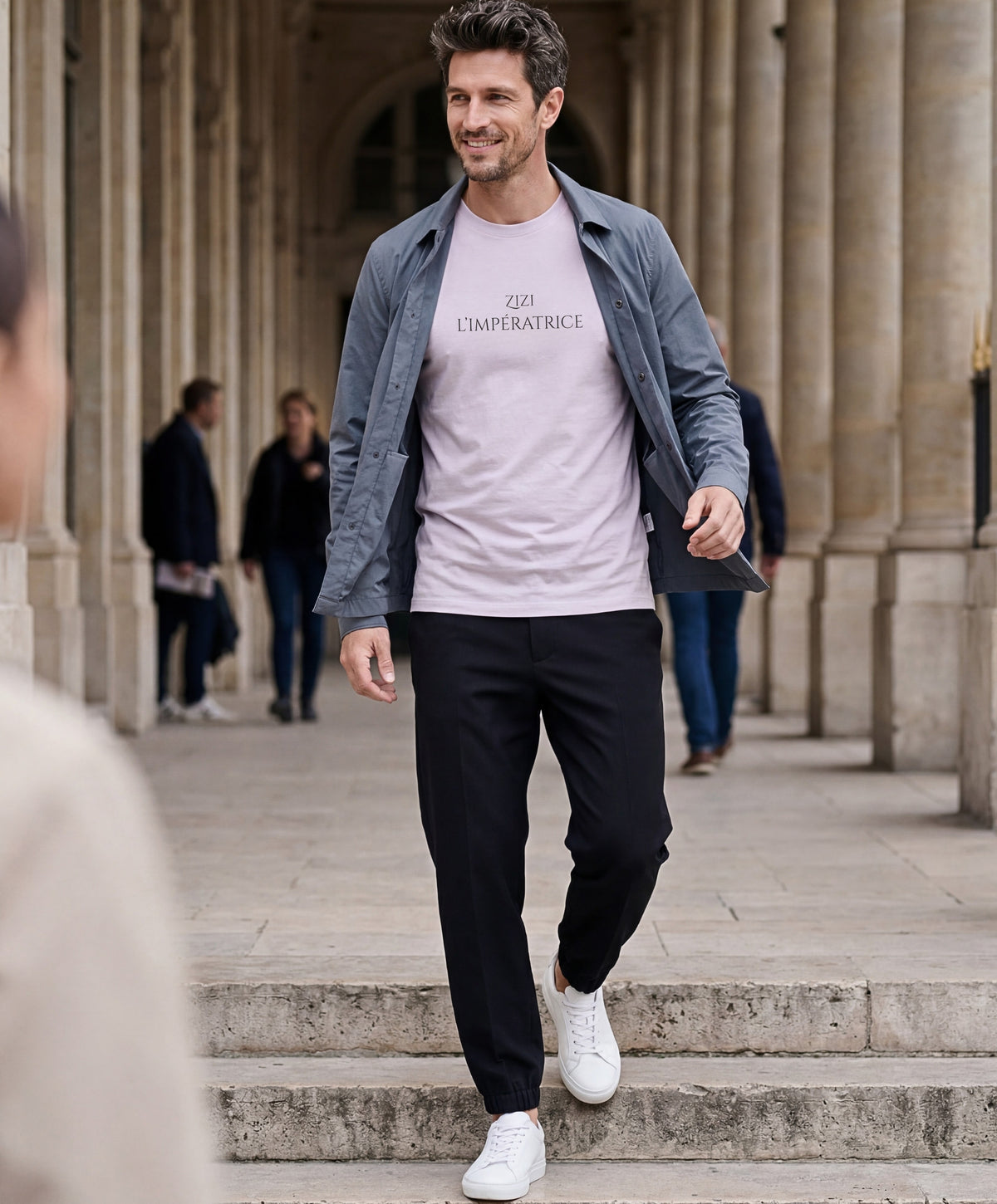 Vue de face d’un homme portant le t-shirt classique lilas « Zizi l’Impératrice » avec texte imprimé noir centré sur la poitrine et logo Salut Fred sur la manche gauche, sous les arcades d’un bâtiment parisien.