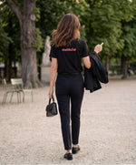 Vue de dos d’une femme portant un t-shirt classique noir Salut Fred avec melèche imprimé au centre du dos dans une teinte melon pastel saumonée, pantalon noir habillé, blazer noir porté à la main et sac noir dans un jardin parisien élégant