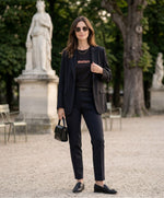 Vue de face d’une femme portant un t-shirt classique noir Salut Fred avec melon imprimé au centre de la poitrine dans une teinte melon pastel saumonée, blazer noir, pantalon noir habillé et sac noir dans un jardin parisien élégant