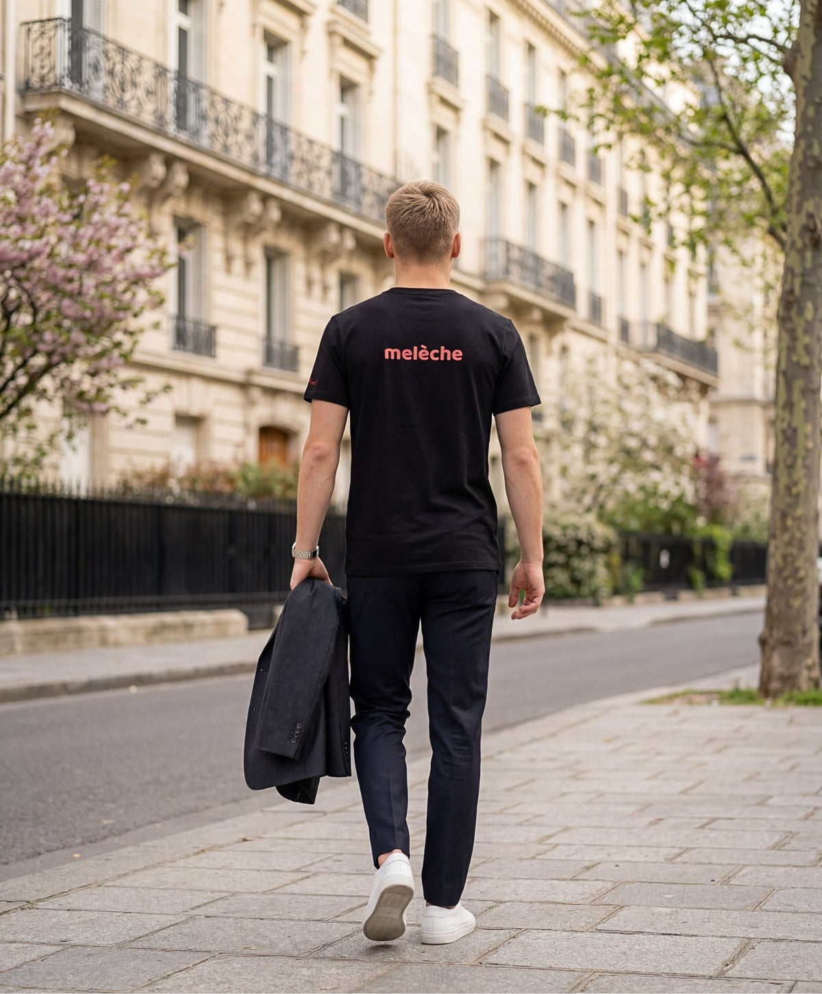 Vue de dos d’un homme portant un t-shirt classique noir Salut Fred avec melèche imprimé au centre du dos dans une teinte melon pastel saumonée, pantalon noir habillé et blazer noir porté à la main dans une rue parisienne élégante