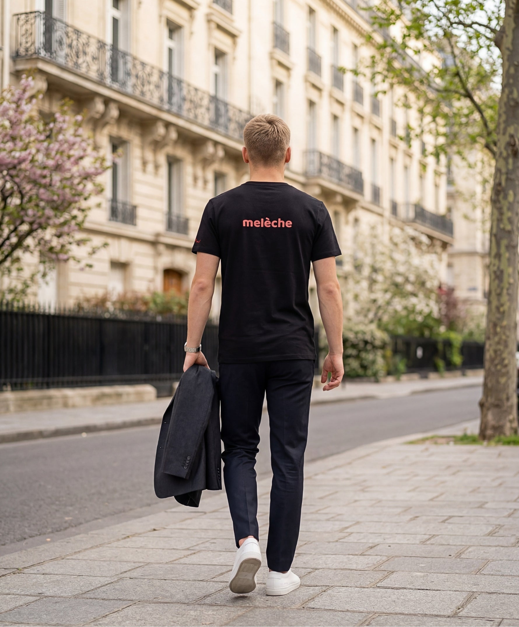 Vue de dos d’un homme portant un t-shirt classique noir Salut Fred avec melèche imprimé au centre du dos dans une teinte melon pastel saumonée, pantalon noir habillé et blazer noir porté à la main dans une rue parisienne élégante