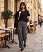 Vue de face d’une femme portant un t-shirt classique noir Salut Fred avec monogramme SF brodé sur la poitrine gauche et broderie Salut Fred sur la manche gauche, pantalon gris graphite habillé, blazer noir, bottines noires et sac noir dans une rue parisienne élégante