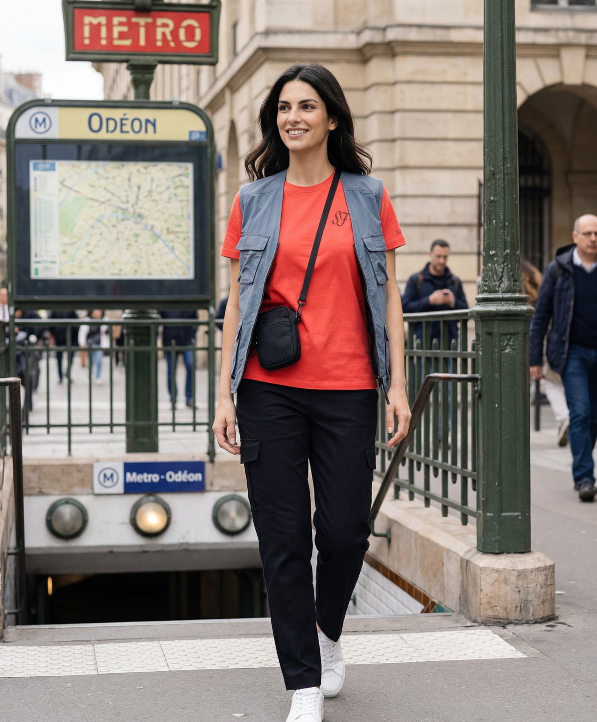 Vue de face d’une femme portant un t-shirt classique orange coquelicot Salut Fred avec monogramme SF brodé sur la poitrine gauche et broderie Salut Fred sur la manche gauche, marchant devant l’entrée du métro Odéon à Paris avec une sacoche bandoulière noire