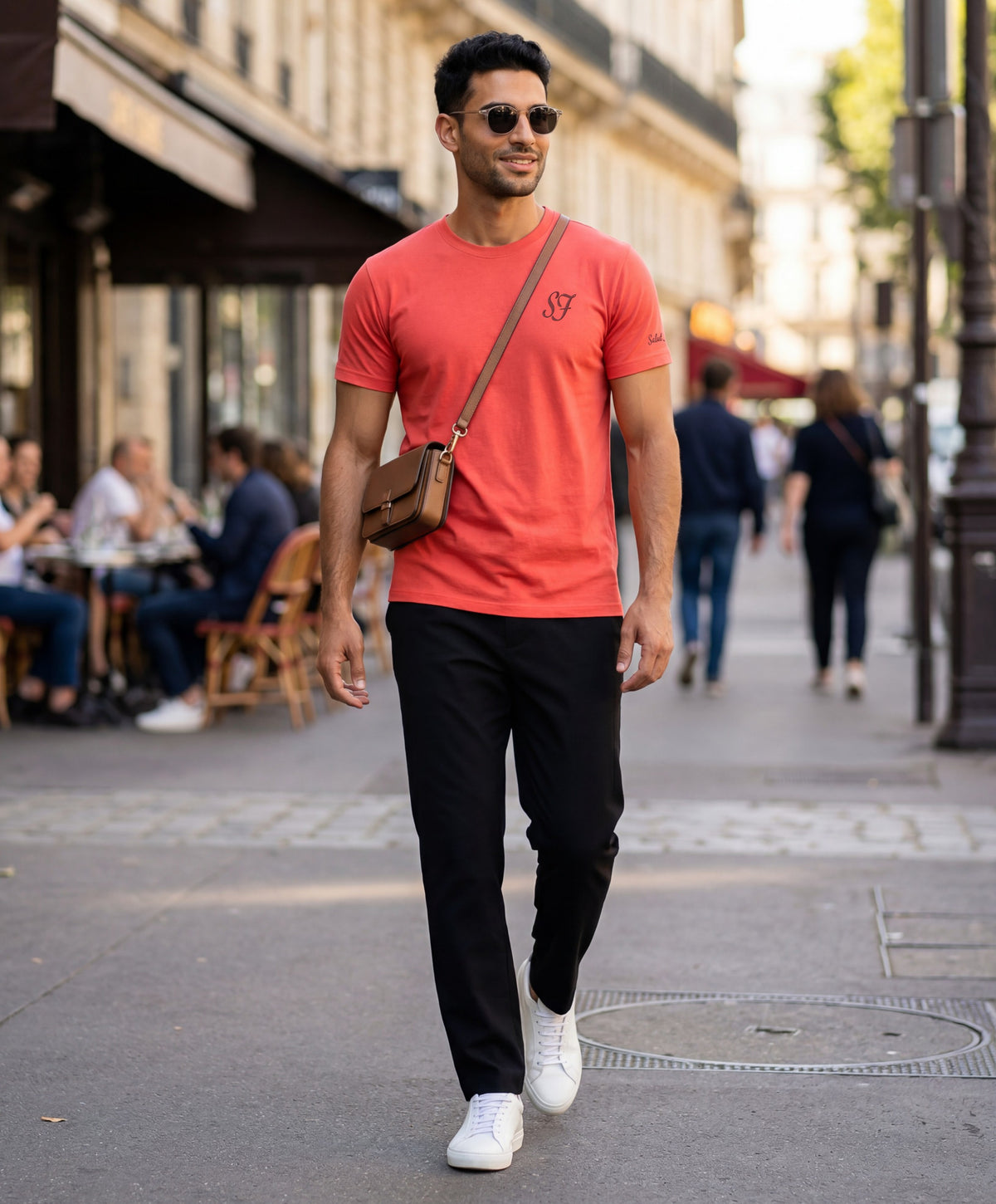 Vue de face d’un homme portant un t-shirt classique orange coquelicot Salut Fred avec monogramme SF brodé sur la poitrine gauche et broderie Salut Fred sur la manche gauche, marchant dans une rue parisienne avec une sacoche bandoulière noire