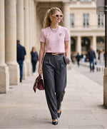 Vue de face d’une femme portant un t-shirt classique rose poudré Salut Fred avec monogramme SF brodé sur la poitrine gauche et broderie Salut Fred sur la manche gauche, pantalon gris graphite habillé, escarpins noirs et sac bordeaux dans une galerie parisienne élégante