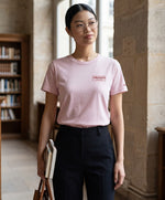 Femme portant un t-shirt classique rose poudré Salut Fred avec broderie « Thérap(e)ute À votre service » sur la poitrine gauche et broderie Salut Fred sur la manche gauche, debout dans une bibliothèque parisienne avec des livres à la main