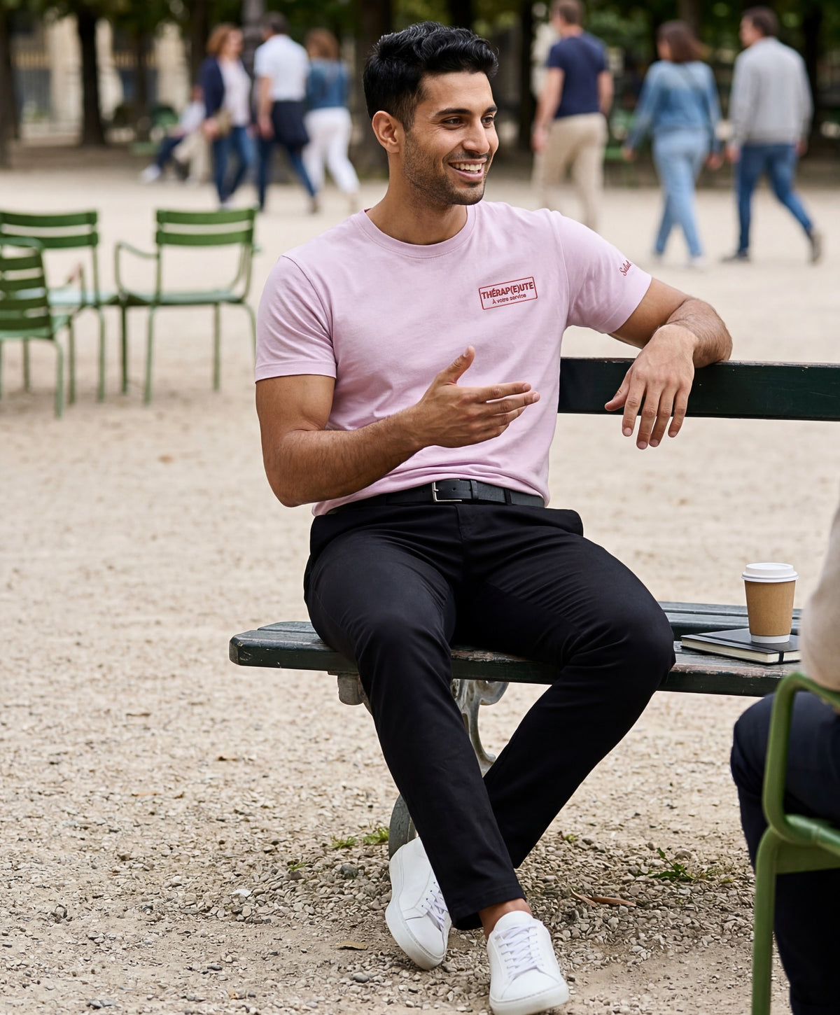 Homme portant un t-shirt classique rose poudré Salut Fred avec broderie « Thérap(e)ute À votre service » sur la poitrine gauche et broderie Salut Fred sur la manche gauche, assis sur un banc dans un jardin parisien