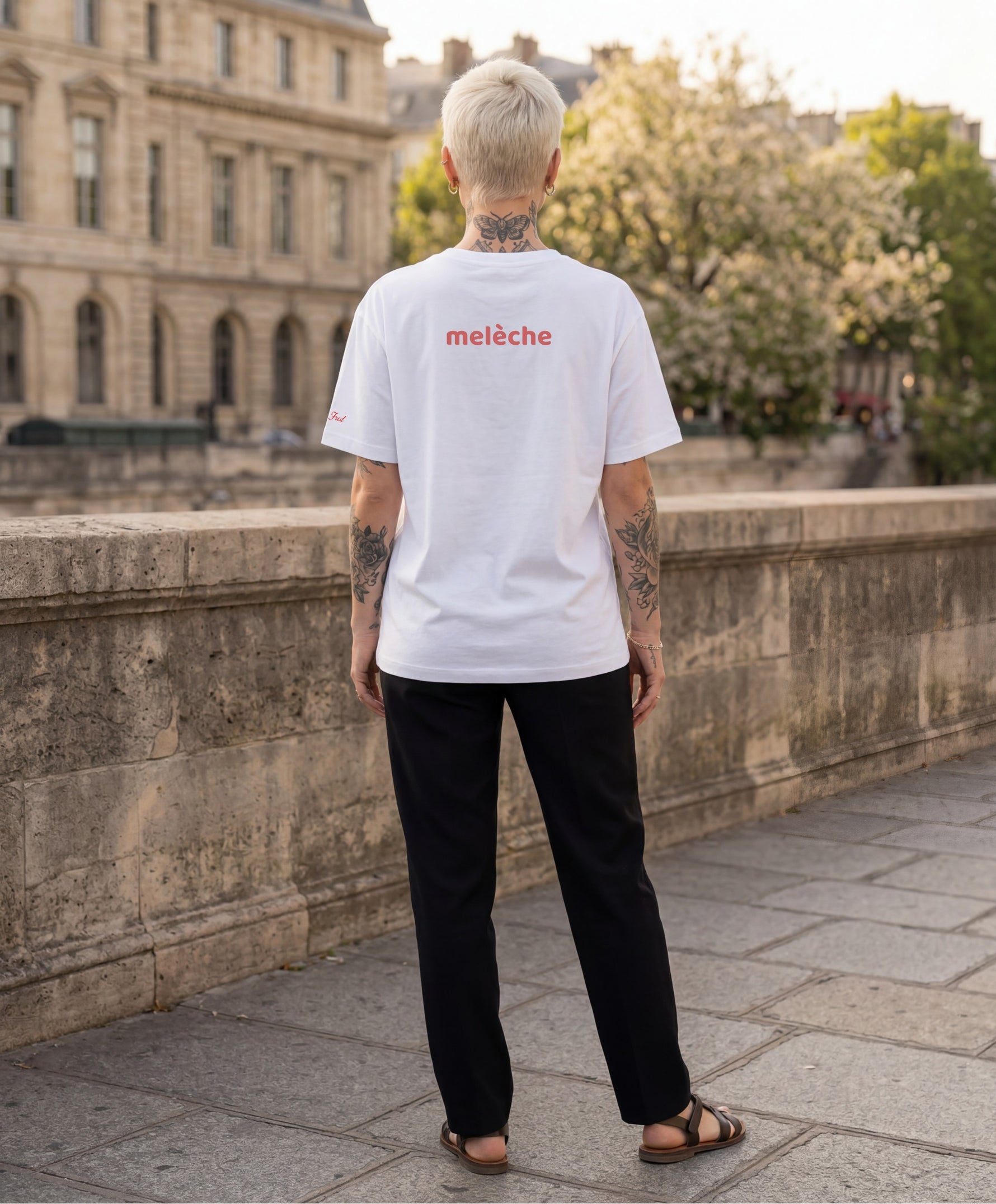 Femme portant le t-shirt oversize blanc « Melon Melèche » avec texte imprimé « melèche » centré dans le dos et logo Salut Fred sur la manche gauche, le long des quais de Seine avec architecture parisienne en arrière-plan.