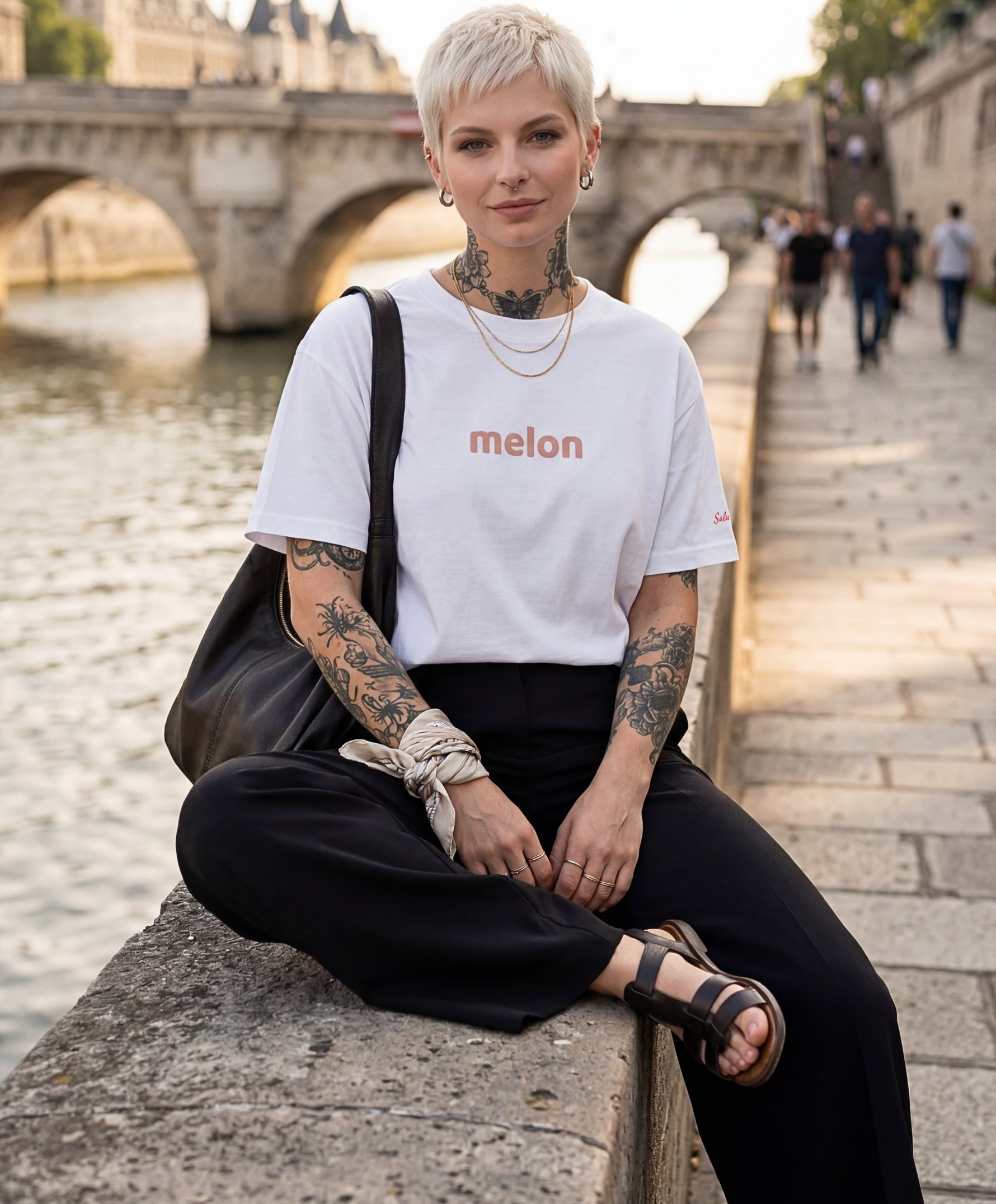Femme portant le t-shirt oversize blanc « Melon Melèche » avec texte imprimé « melon » centré sur la poitrine et logo Salut Fred sur la manche gauche, assise le long des quais de Seine avec un pont parisien en arrière-plan.