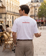 Homme portant le t-shirt oversize blanc « Melon Melèche » avec texte imprimé « melèche » centré dans le dos et logo Salut Fred sur la manche gauche, dans une rue parisienne bordée de cafés et d’immeubles haussmanniens.